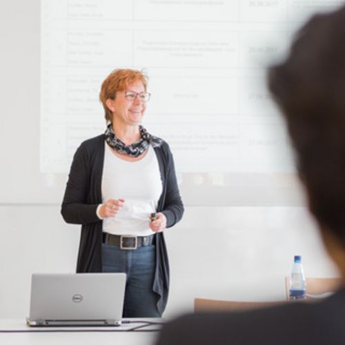 Eine Dozentin steht vor einer Wand, auf die eine Pr&auml;sentation projiziert wird. Sie spricht zu Studierenden in einem Seminarraum.