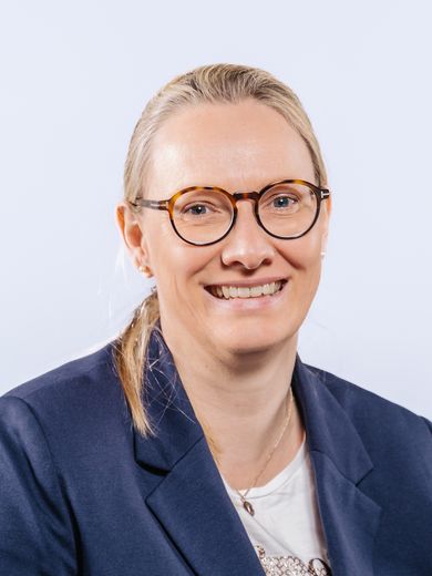 Auf dem Bild ist Esther Blömer zu sehen. Sie hat ihr blondes Haar zu einem flachen Zopf gebunden. Sie trägt eine runde Brille und einen dunkelblauen Blazer. 