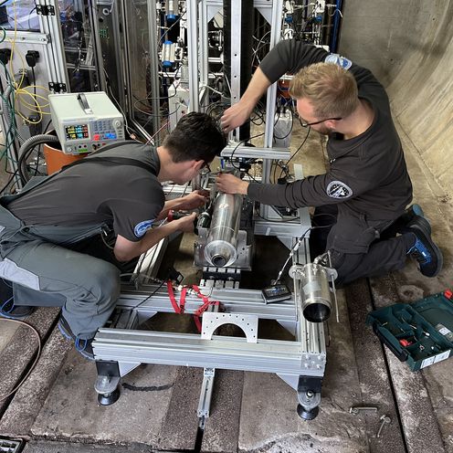 Auf dem Bild sind die Vorbereitungen des Triebwerktests zusehen. Zwei Studenten sitzen vor dem Triebwerk in einer gro&szlig;en Betonr&ouml;hre und schrauben an dem Triebwerk herum. 