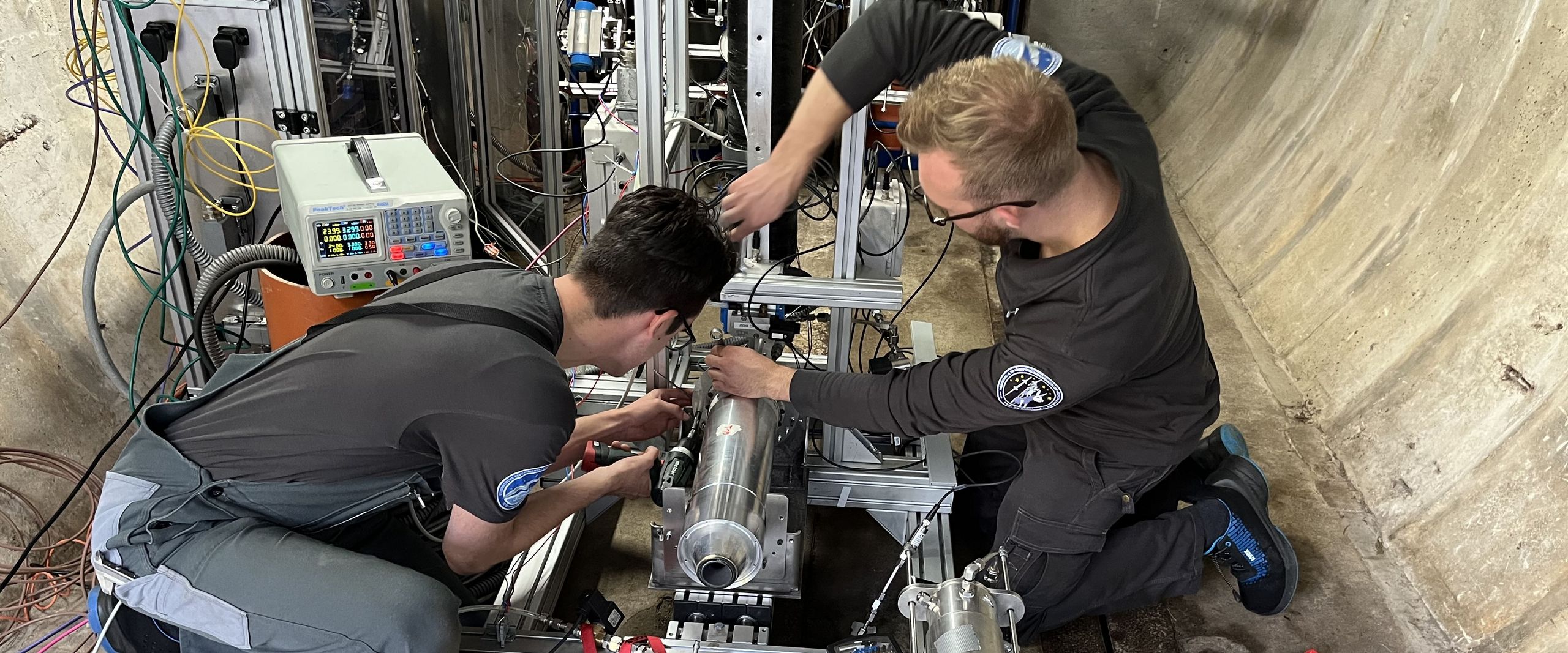 Auf dem Bild sind die Vorbereitungen des Triebwerktests zusehen. Zwei Studenten sitzen vor dem Triebwerk in einer großen Betonröhre und schrauben an dem Triebwerk herum. 