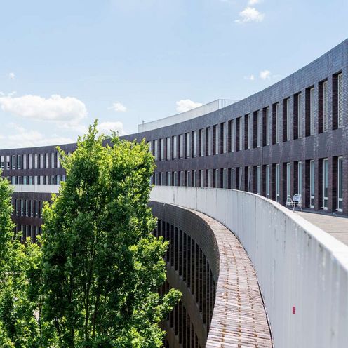 Auf dem Bild ist eine Au&szlig;enansicht des ZIMT-Geb&auml;udes zu sehen. Die Form des Geb&auml;udes &auml;hnelt einem Halbkreis aus dunkelroten Backsteinen. Es sieht sehr modern aus. In der Rundung des Geb&auml;udes wachsen B&auml;ume. 