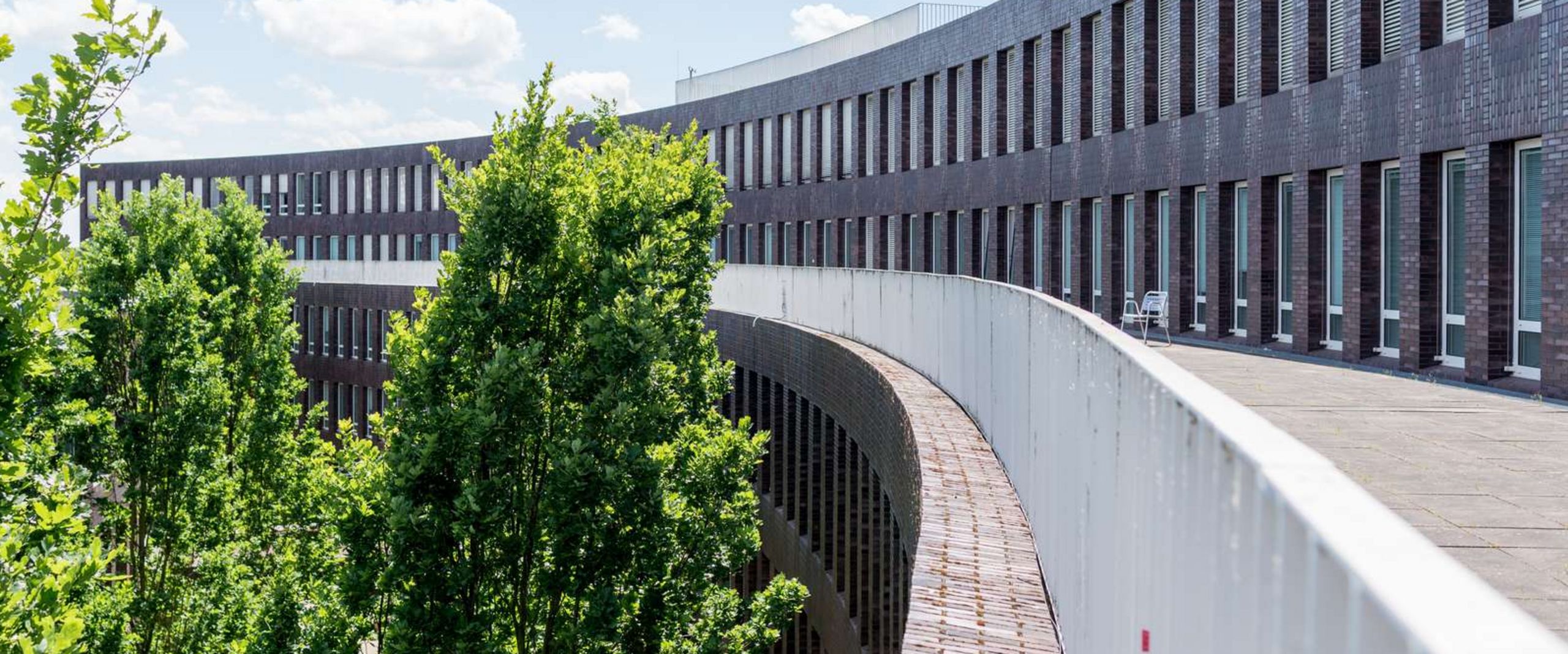 ZIMT-Gebäude Auf dem Bild ist eine Außenansicht des ZIMT-Gebäudes zu sehen. Die Form des Gebäudes ähnelt einem Halbkreis aus dunkelroten Backsteinen. Es sieht sehr modern aus. In der Rundung des Gebäudes wachsen Bäume.