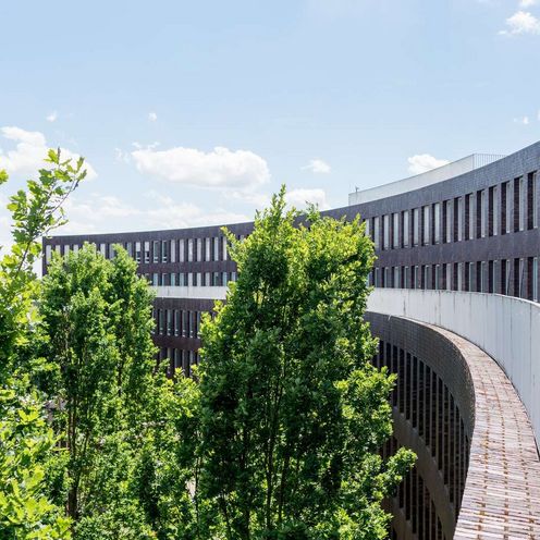 Auf dem Bild ist eine Au&szlig;enansicht des ZIMT-Geb&auml;udes zu sehen. Die Form des Geb&auml;udes &auml;hnelt einem Halbkreis aus dunkelroten Backsteinen. Es sieht sehr modern aus. In der Rundung des Geb&auml;udes wachsen B&auml;ume. 