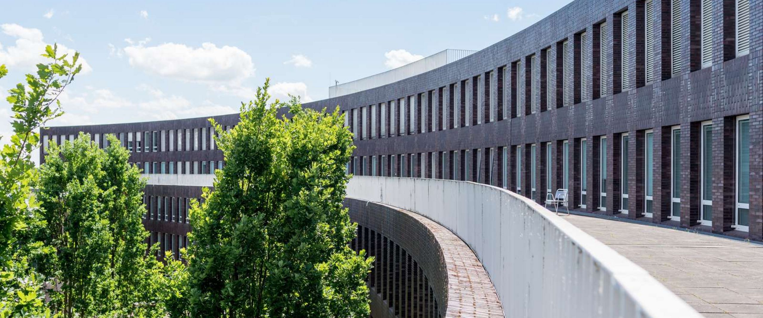 ZIMT-Gebäude Auf dem Bild ist eine Außenansicht des ZIMT-Gebäudes zu sehen. Die Form des Gebäudes ähnelt einem Halbkreis aus dunkelroten Backsteinen. Es sieht sehr modern aus. In der Rundung des Gebäudes wachsen Bäume.