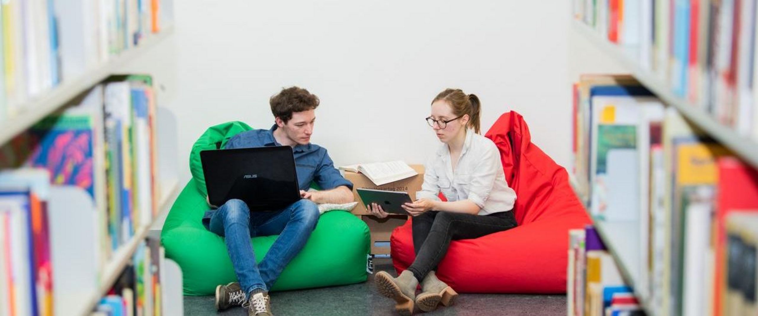 Eine Studentin und ein Student sitzen mit Laptops in Sitzsäcken in der Bibliothek , im Vordergrund Bücherregale