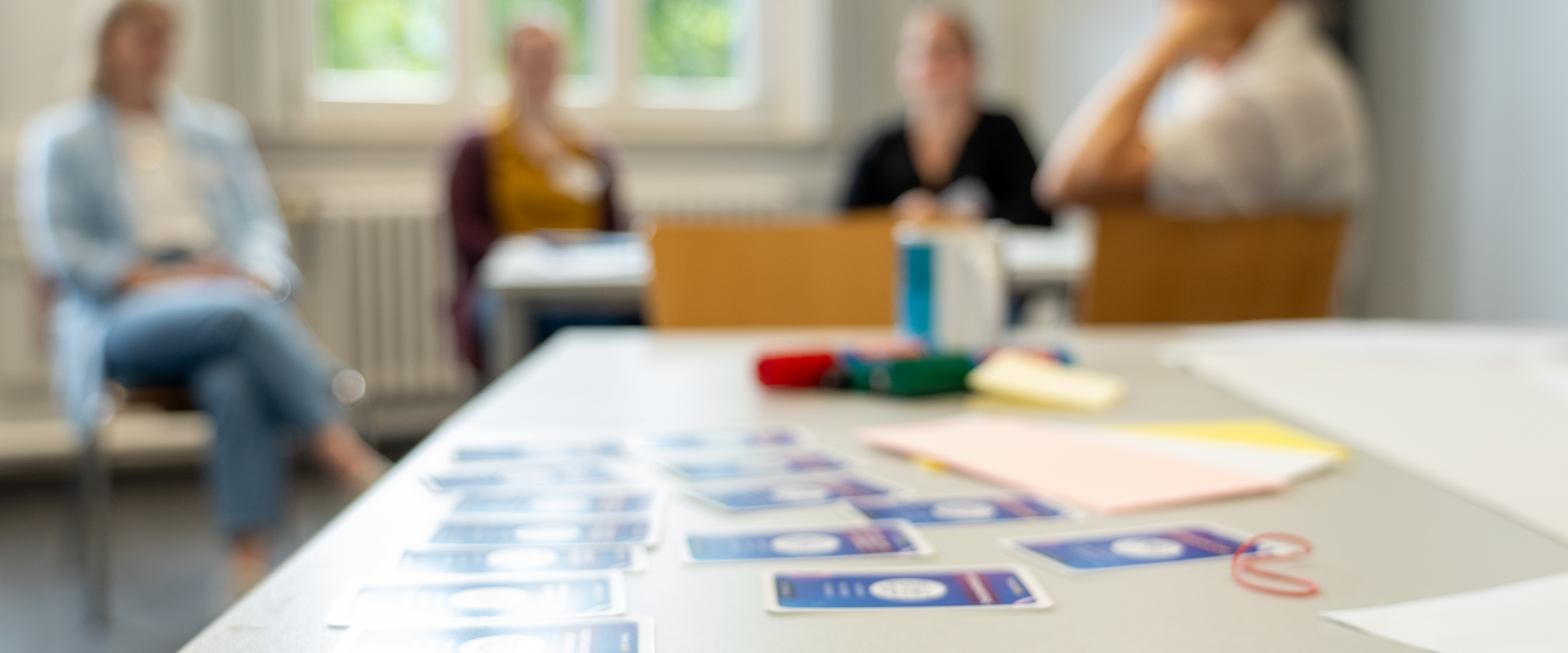 Gesprächssituation in einem Seminarraum: Vier Personen sitzen zusammen, auf dem Tisch liegen Moderationskarten und Materialien.