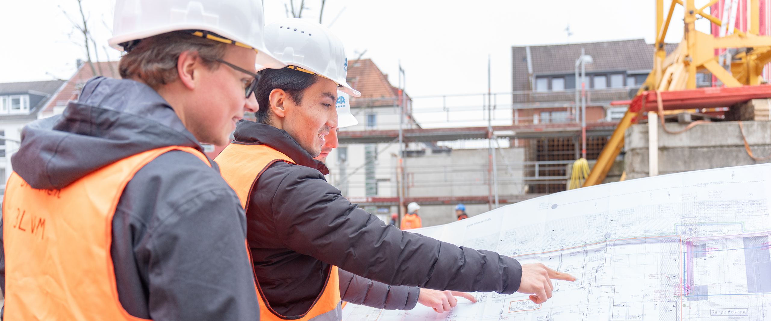 Baustellenplanung Auf dem Bild sind zwei junge Männer mit Warnweste und Schutzhelm zu sehen. Sie stehen auf einer Baustelle und halten einen großen Bauplan in den Händen.