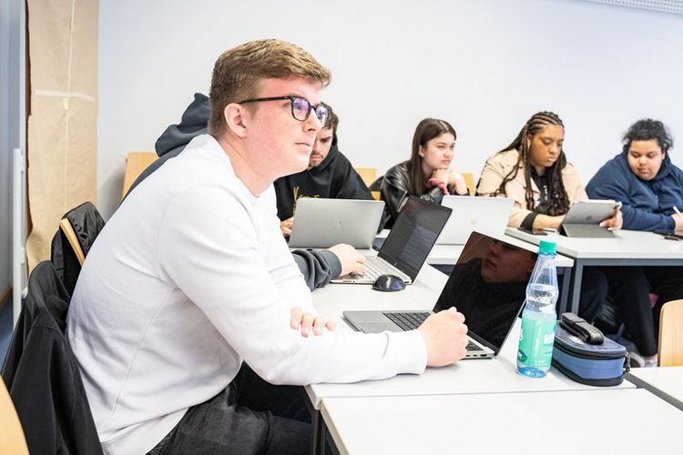 Studierende sitzen über Eck an Tischen in einem Seminarraum. Sie schauen teils nach vorn, teils auf ihre Notizen oder Notebooks, die vor ihnen platziert sind.