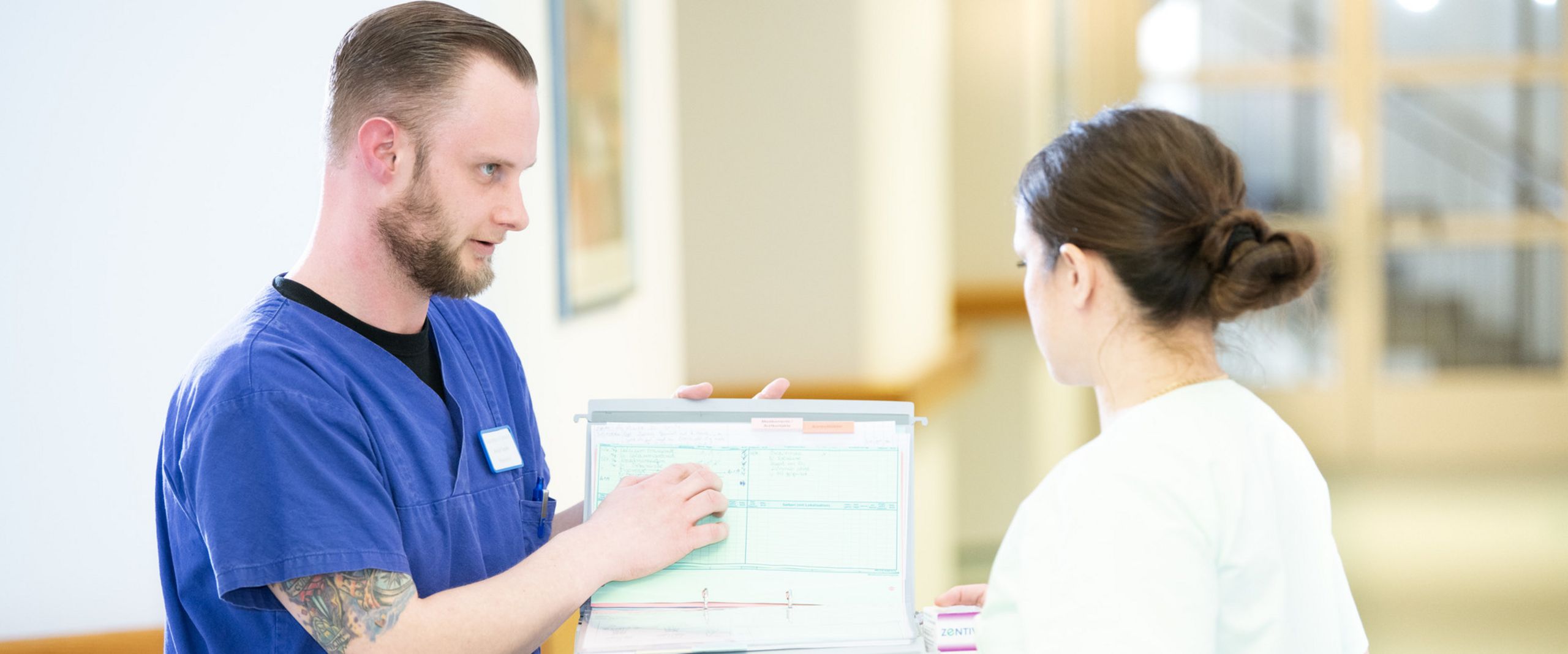 Ein Pfleger und eine Pflegerin besprechen sich an einem Dokumentenwagen auf dem Flur eines Seniorenheimes.