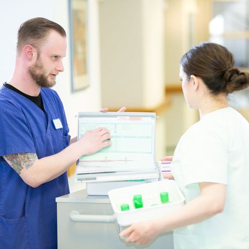 Ein Pfleger und eine Pflegerin besprechen sich an einem Dokumentenwagen auf dem Flur eines Seniorenheimes.