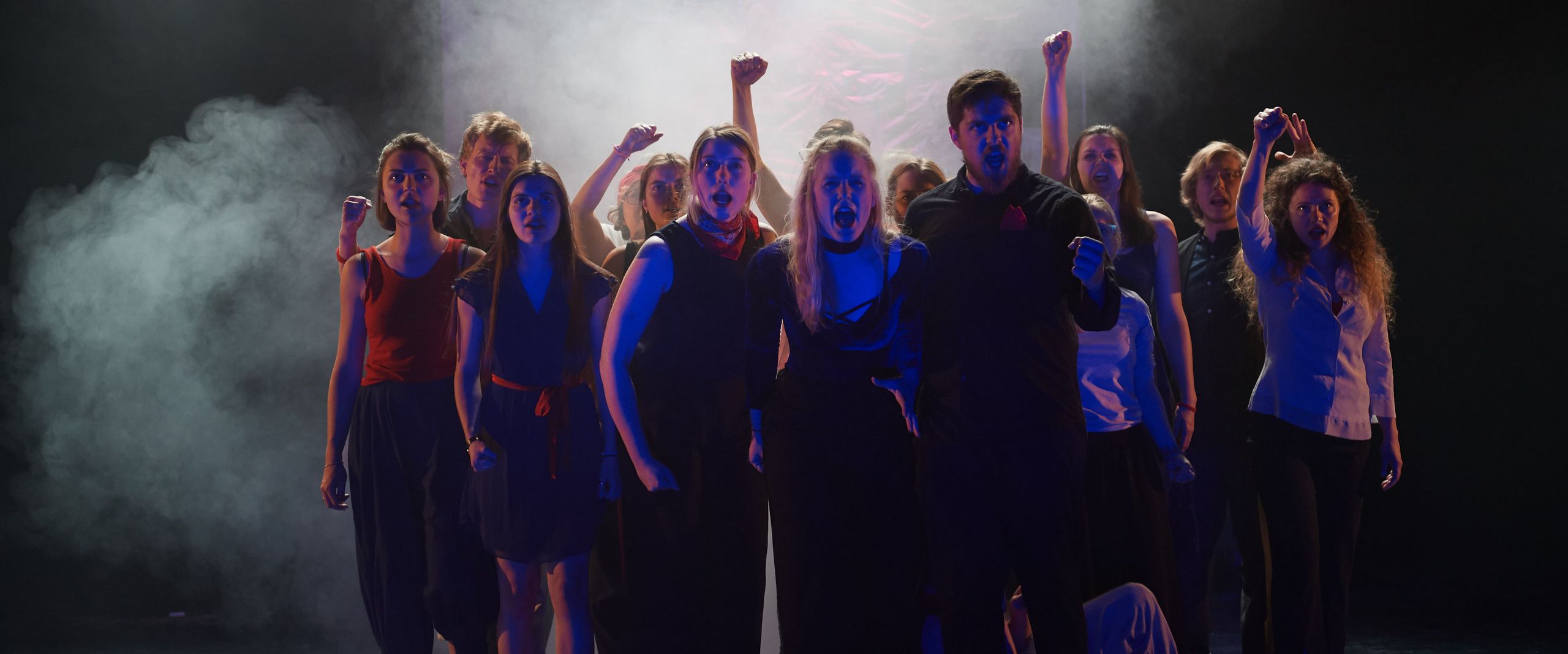 Dunkel gekleidete Menschen stehen auf einer Bühne. Es ist dunkel und Nebel umgibt sie.