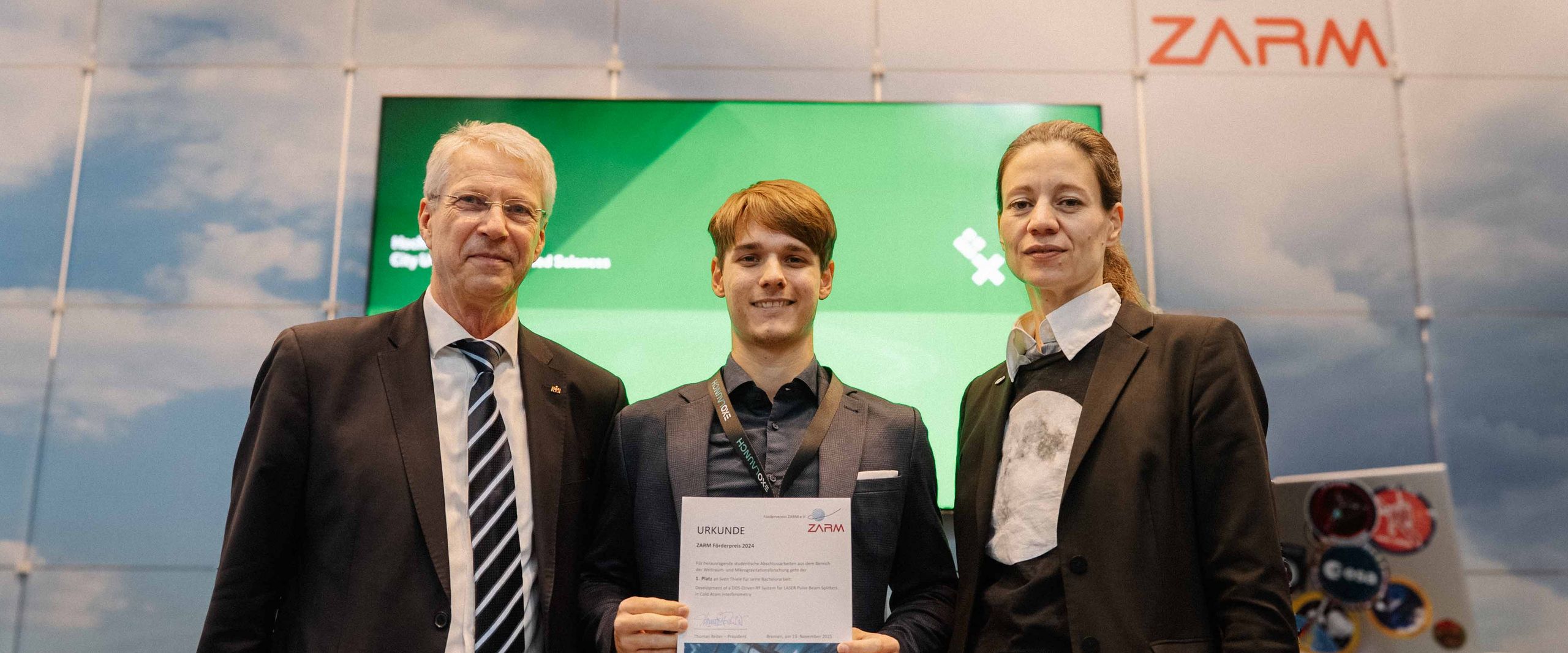 Gruppenbild mit zwei M&auml;nnern und einer Frau. Der Mann in der Mitte h&auml;lt eine Urkunde.