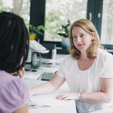 Eine Frau sitzt an einem Schreibtisch und hält Unterlagen in der Hand. Sie blickt zu einem Mädchen, welche nur von hinten zu sehen ist.