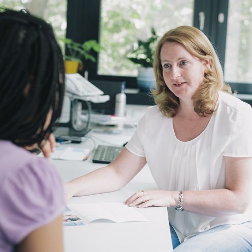 Eine Frau sitzt an einem Schreibtisch und h&auml;lt Unterlagen in der Hand. Sie blickt zu einem M&auml;dchen, welche nur von hinten zu sehen ist.