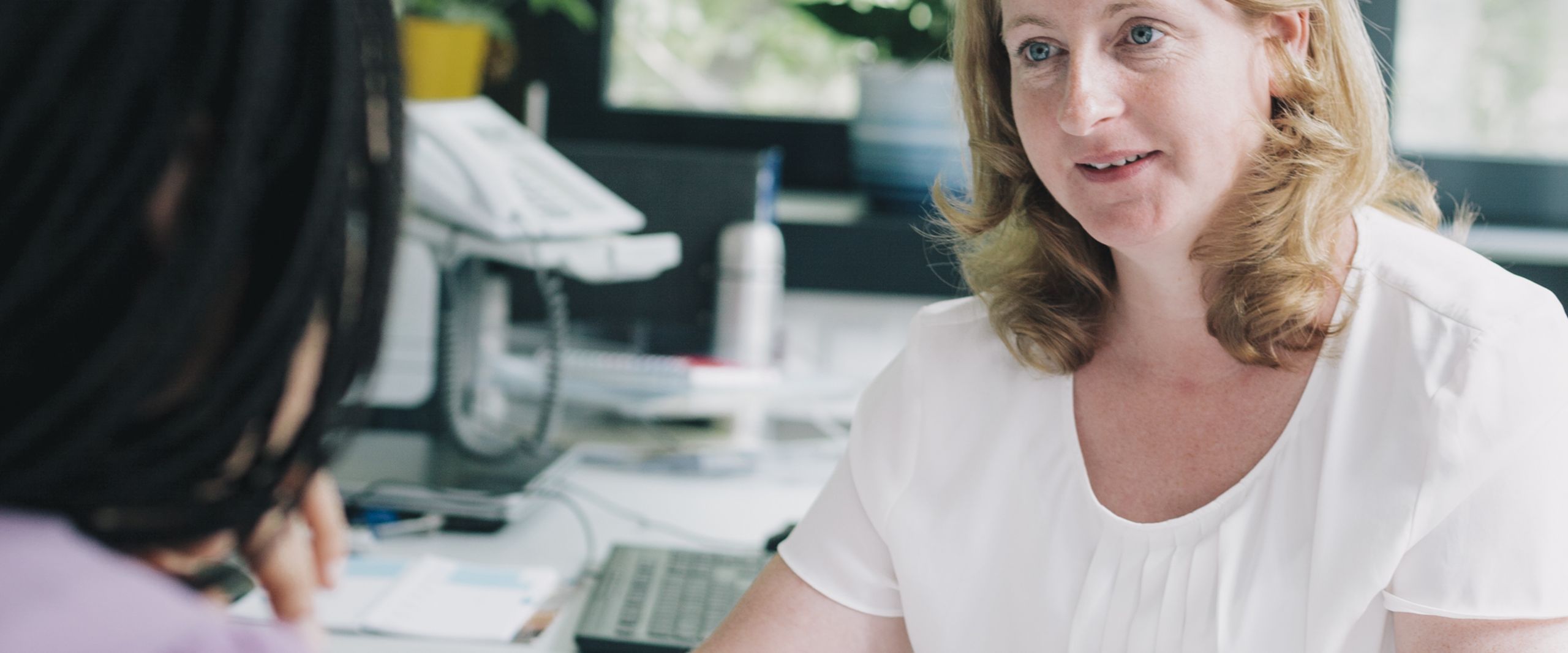 Eine Frau sitzt an einem Schreibtisch und h&auml;lt Unterlagen in der Hand. Sie blickt zu einem M&auml;dchen, welche nur von hinten zu sehen ist.