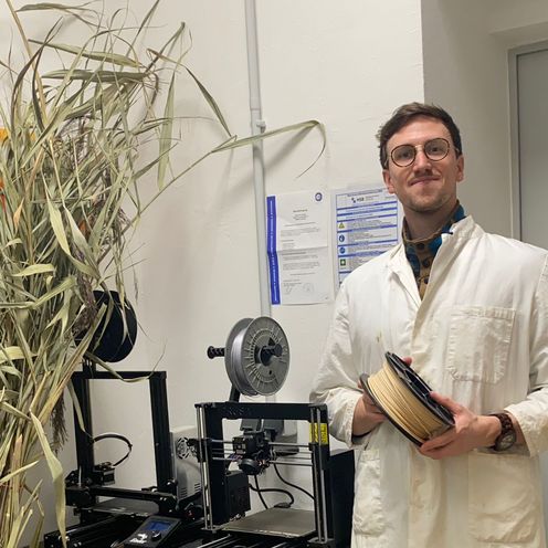 Jonas Baumann posiert im Laborkittel vor einem 3D-Drucker