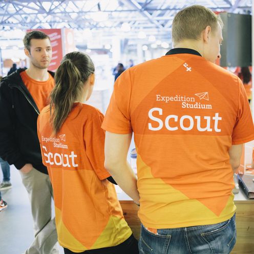 Drei junge M&auml;nner und Frauen tragen organene T-Shirts. Sie sind mit dem R&uuml;cken zu sehen. Auf ihren T-Shirts steht: Scouts, Experiment Studium.