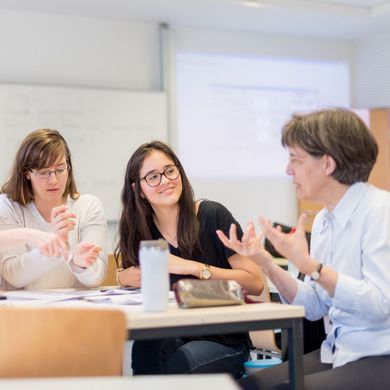 Professorin Martina Röhrich sitzt am Tisch in einem Seminarraum und diskutiert mit einer kleinen Gruppe Studierender.
