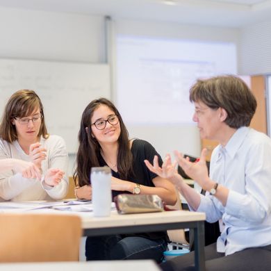 Professorin Martina Röhrich sitzt am Tisch in einem Seminarraum und diskutiert mit einer kleinen Gruppe Studierender.