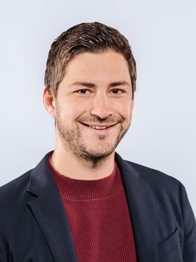 Auf dem Bild ist Jonas Unterholzner zusehen. Er hat braune Haare und einen Dreitagebart. Er trägt einen dunkel roten Pullover unter einem dunkel blauen Jacket. 