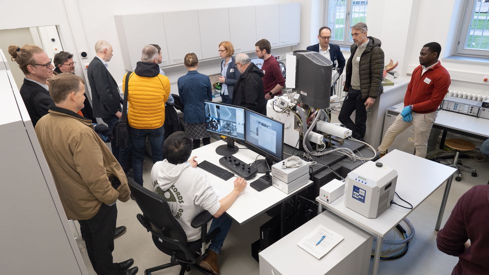 Labor mit Ger&auml;ten. Zahlreiche M&auml;nner und Frauen schauen sich um.