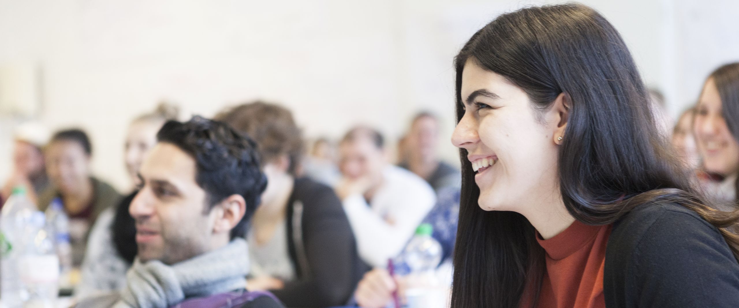 Student mit kurzen dunklen, leicht lockigen Haaren und Studentin mit langen, dunklen Haaren, beide sitzend vor einer Gruppe weiterer Studierender