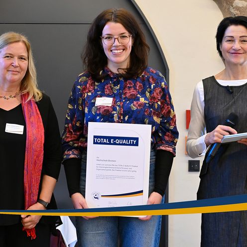TOTAL E-QUALITY Prädikat 2026 Drei Frauen stehen nebenenander. Luise Kilian in der Mitte hält die Urkunde des TOTAL E-QUALITY Prädikats in der Hand. Alle lächeln.