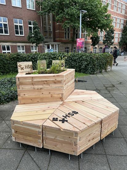 Holzmodule in Wabenform mit Sitzflächen und integrierten Pflanzkästen auf dem Mensavorplatz der Hochschule Bremen. Auf den Modulen ist das Logo ‚HSB‘ zu sehen.