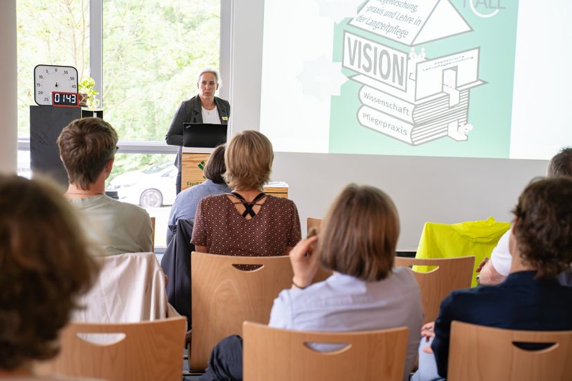 Auf dem Bild ist eine Frau zusehen, die vor einer Gruppe sitzender Menschen eine Präsentation hält. Sie hat lange weiße Haare und trägt einen Blazer. 