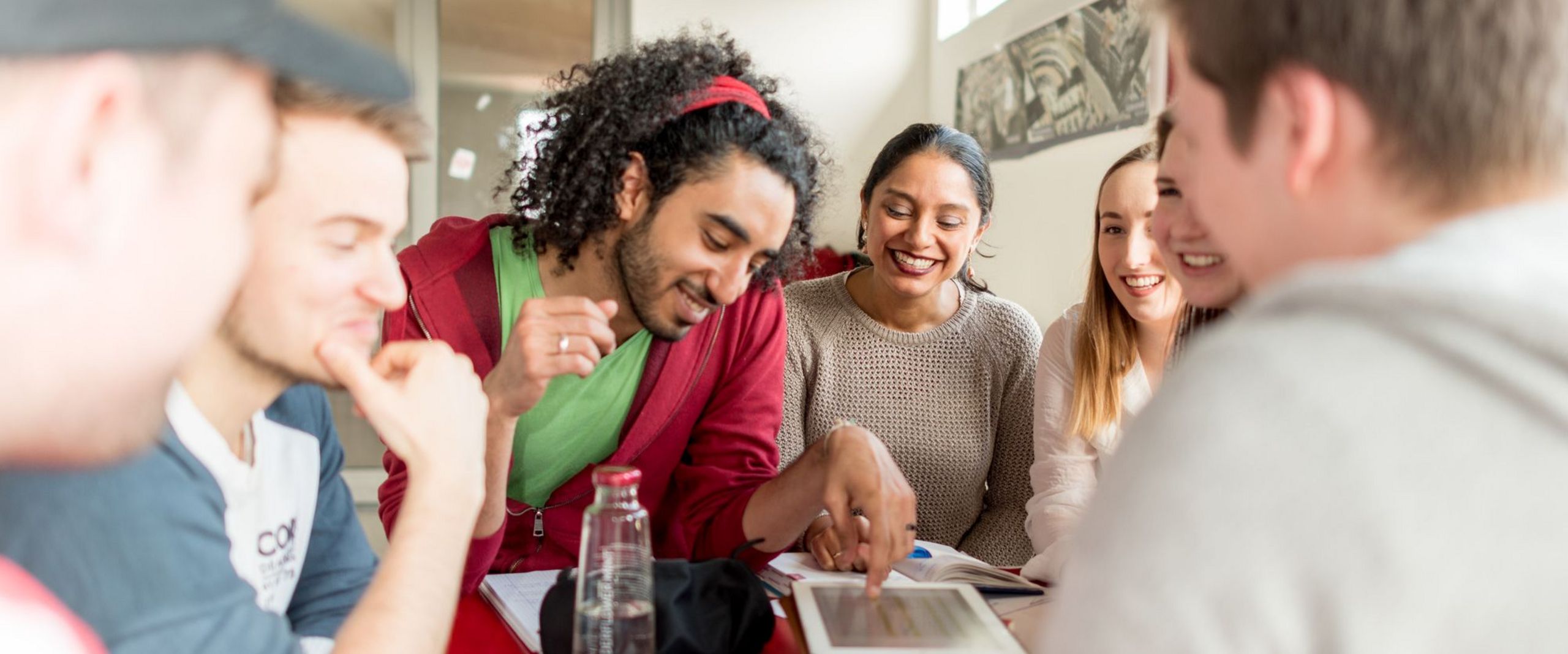 Eine Gruppe von Studierenden sitzt an einem Tisch. Sie lachen und blicken teilweise auf ein Tablet vor ihnen auf dem Tisch.