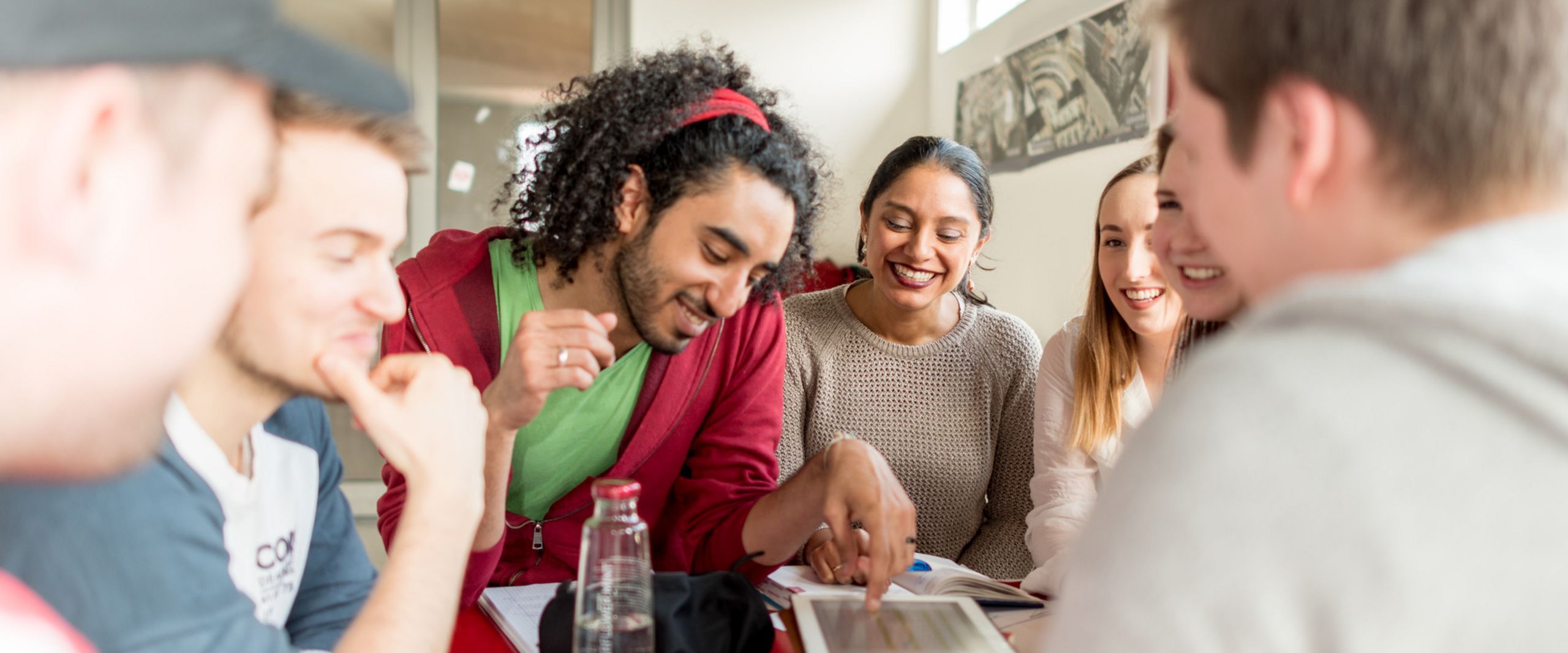 Studierende Eine Gruppe von Studierenden sitzt an einem Tisch. Sie lachen und blicken teilweise auf ein Tablet vor ihnen auf dem Tisch.