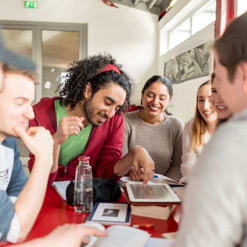 Eine Gruppe von Studierenden sitzt an einem Tisch. Sie lachen und blicken teilweise auf ein Tablet vor ihnen auf dem Tisch.