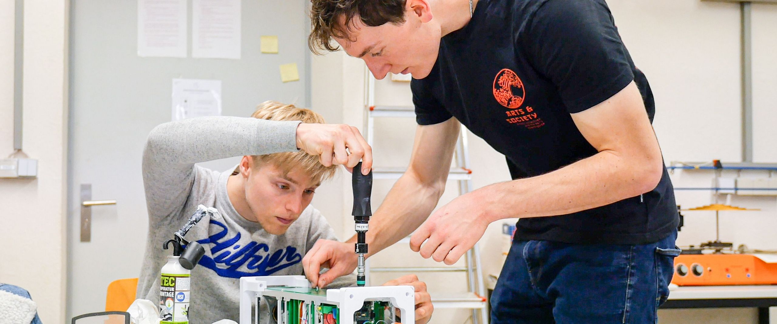 Zwei Studierende schrauben an einem technischen Gerät auf einem Tisch.