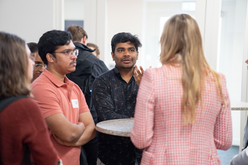 Menschen stehen im Kreis um einen Stehtisch herum und unterhalten sich. Im Hintergrund sind weitere Menschen.
