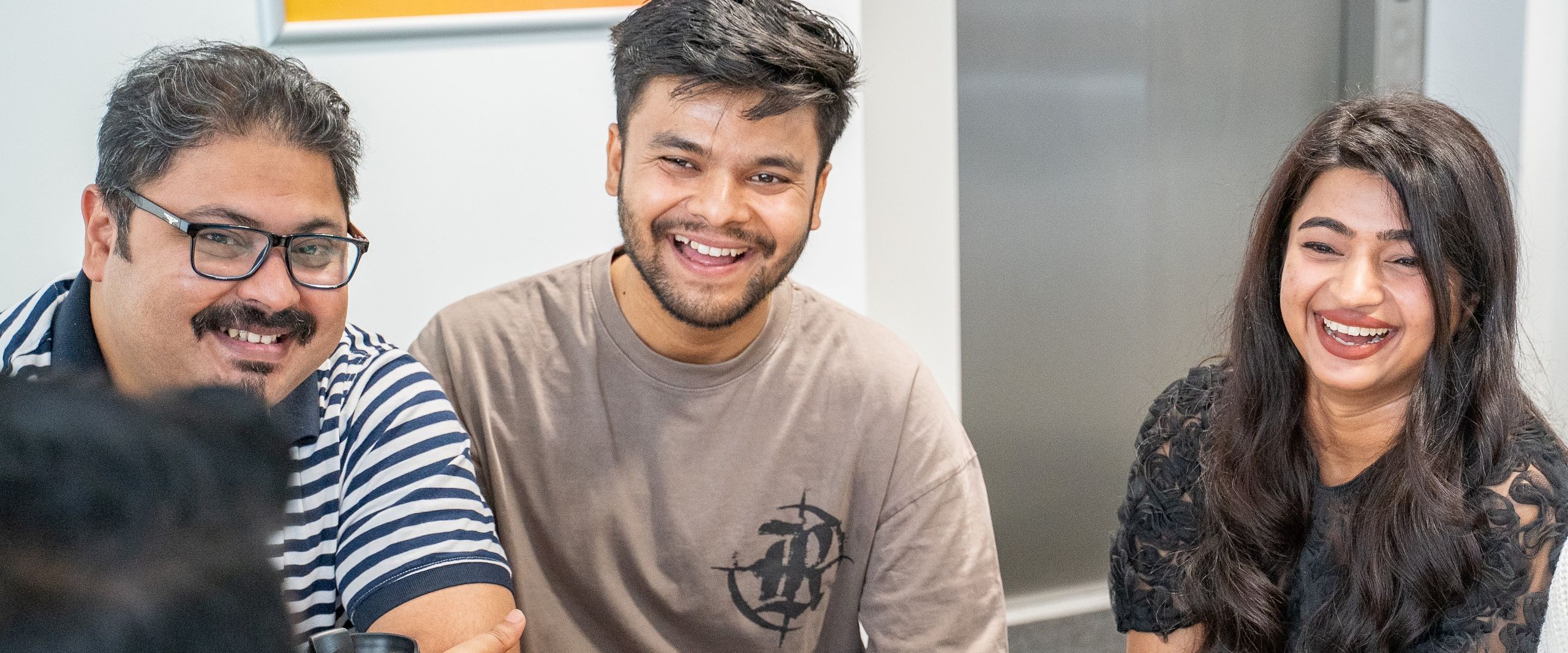 3 lachende Studierende an einem Tisch, eine junge Frau, 2 junge M&auml;nner