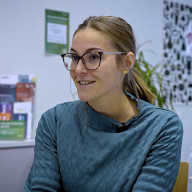 Auf dem Bild ist eine Frau zusehen. Sie sitzt an einem Tisch. Sie hat ihre blonden Haare zu einem Zopf zusammengebunden und tr&auml;gt einen blauen Pullover.