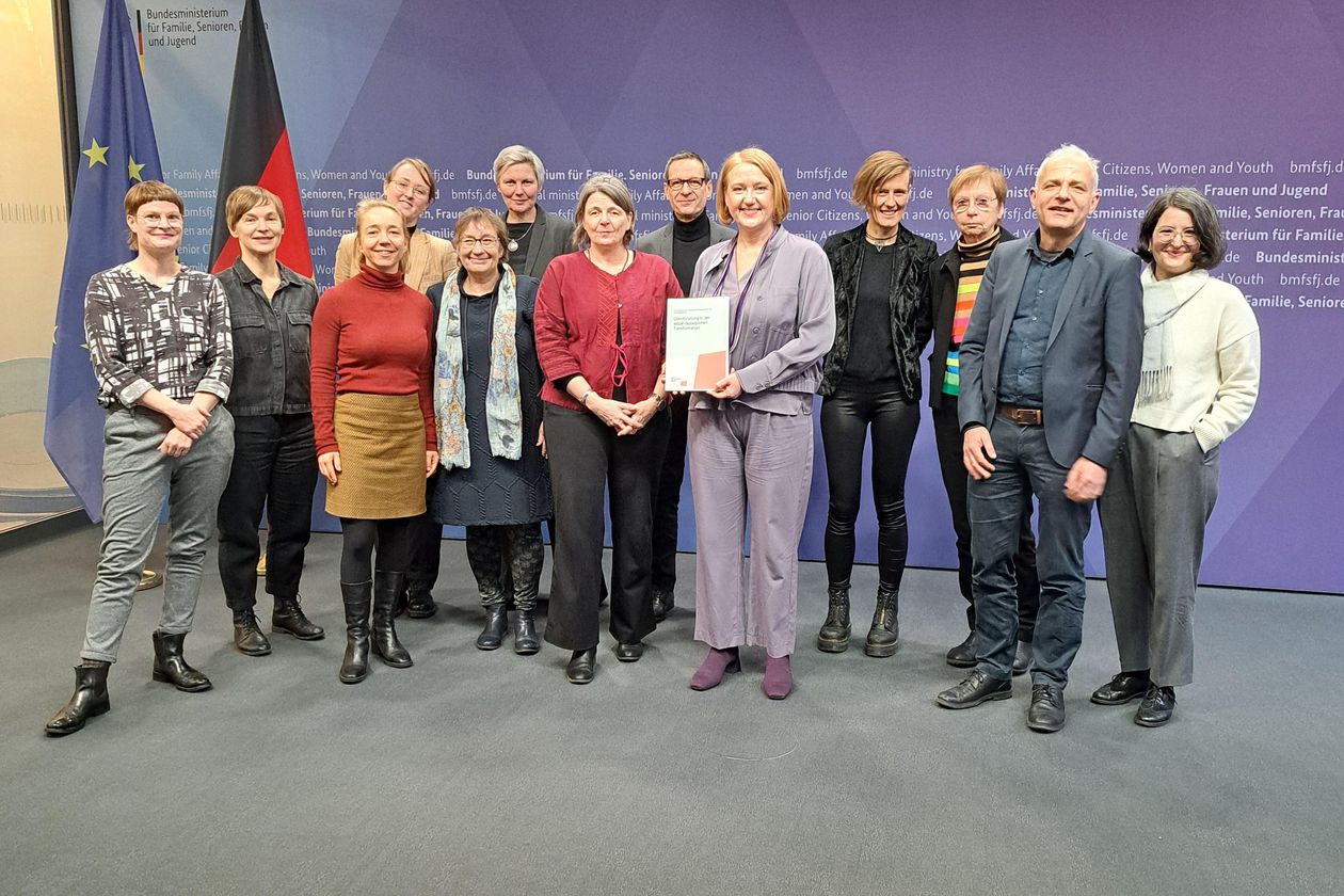 Auf dem Bild ist eine Gruppe von Leuten zusehen. Sie stehen in zwei Reihen und schauen in die Kamera. Eine Frau in der Mitte mit einem lila Hosenanzug hat einen ausgedruckten Bericht in der Hand. 