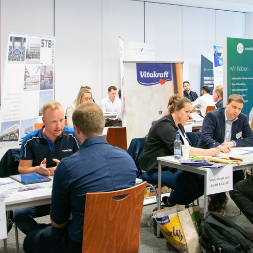 M&auml;nner und Frauen sitzen in einem Raum an Tischen und sind im Gespr&auml;ch. Um sie herum stehen Roll-ups.