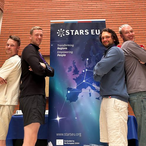 Drei M&auml;nner stehen links und rechts von einem Roll-up und l&auml;cheln in die Kamera.