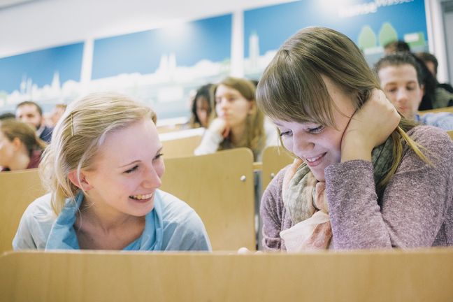 Zwei junge Frauen sitzen einander zugewandt und lächelnd im Hörsaal.