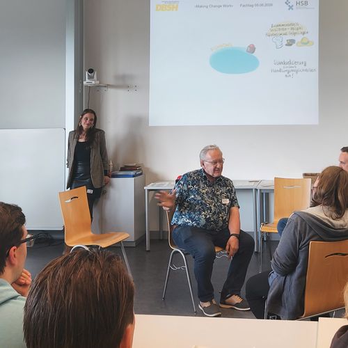 Mehrere Menschen sitzen im Stuhlkreis und reden miteinander. Andere sitzen an Seminartischen und h&ouml;reren zu.