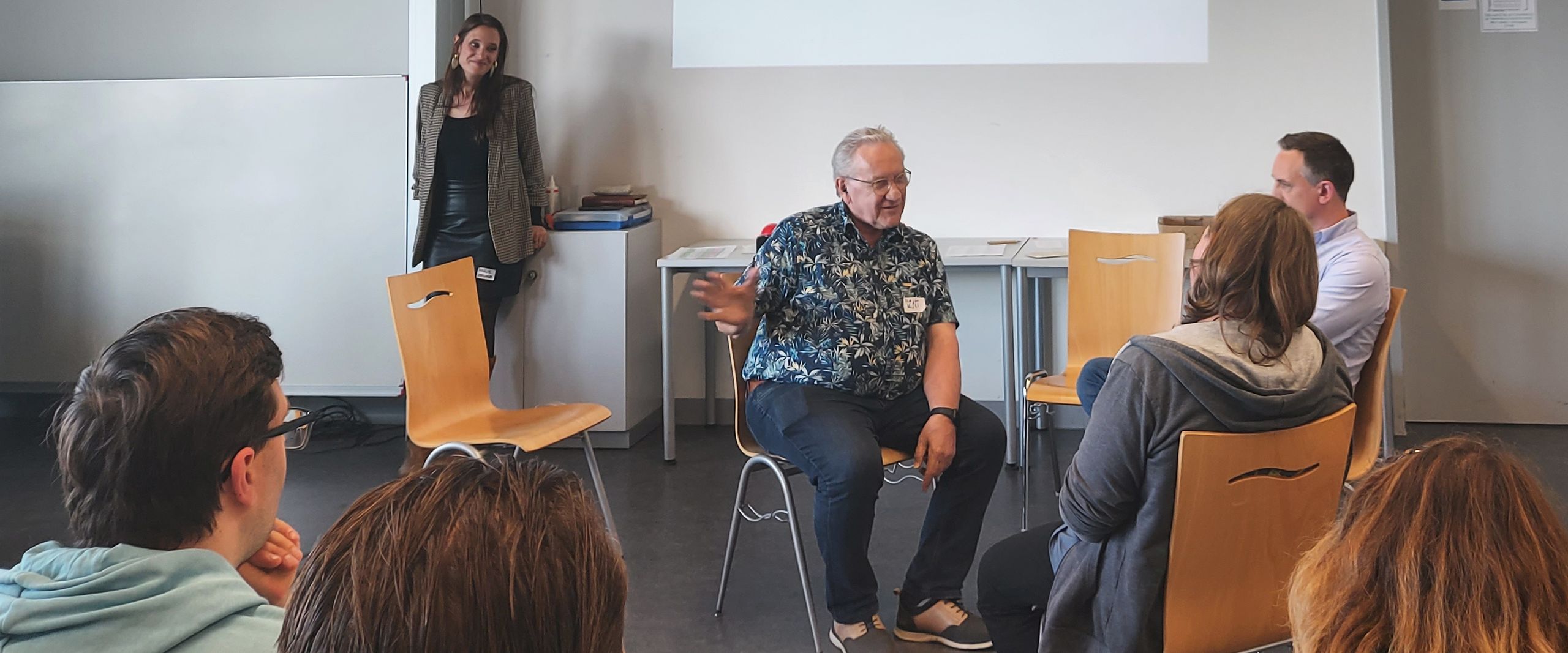 Mehrere Menschen sitzen im Stuhlkreis und reden miteinander. Andere sitzen an Seminartischen und h&ouml;reren zu.