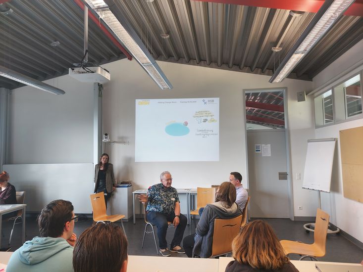 Mehrere Menschen sitzen im Stuhlkreis und reden miteinander. Andere sitzen an Seminartischen und h&ouml;reren zu.