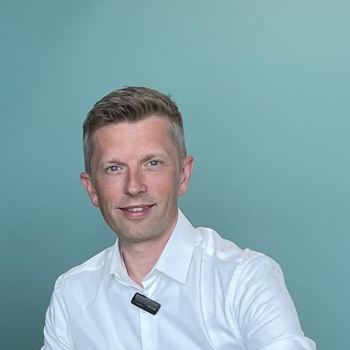 Zwei M&auml;nner sitzen am Tisch und alchen in die Kamera. Rechts von ihnen steht ein Laptop.