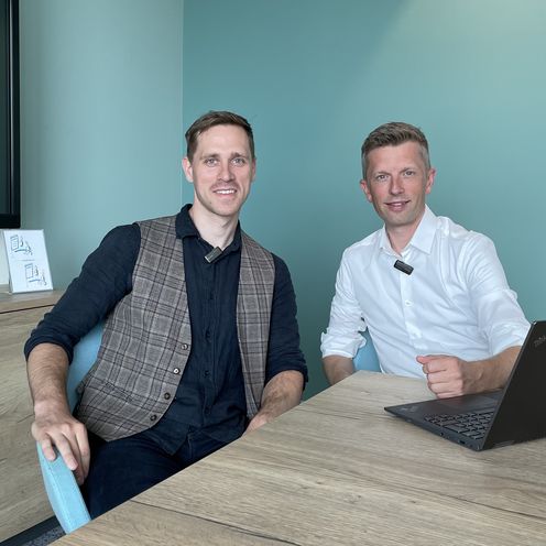 Zwei M&auml;nner sitzen am Tisch und alchen in die Kamera. Rechts von ihnen steht ein Laptop.