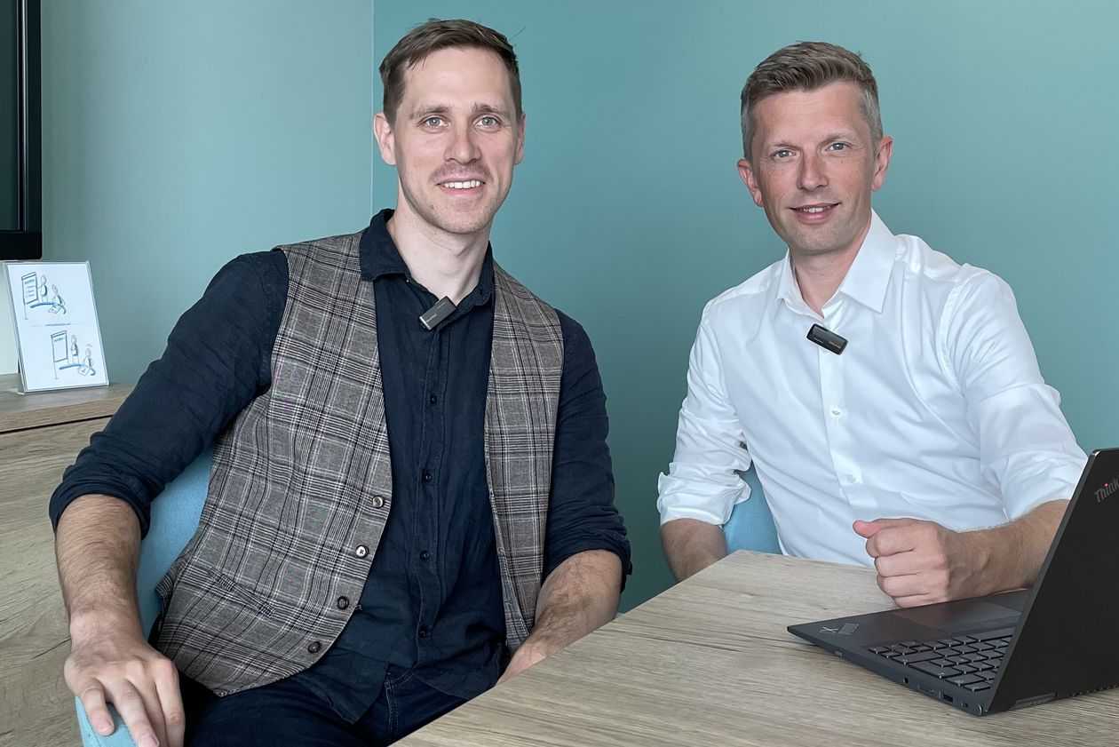 Zwei M&auml;nner sitzen am Tisch und alchen in die Kamera. Rechts von ihnen steht ein Laptop.