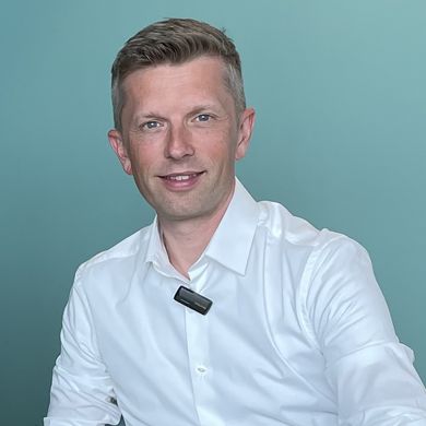 Zwei M&auml;nner sitzen am Tisch und alchen in die Kamera. Rechts von ihnen steht ein Laptop.