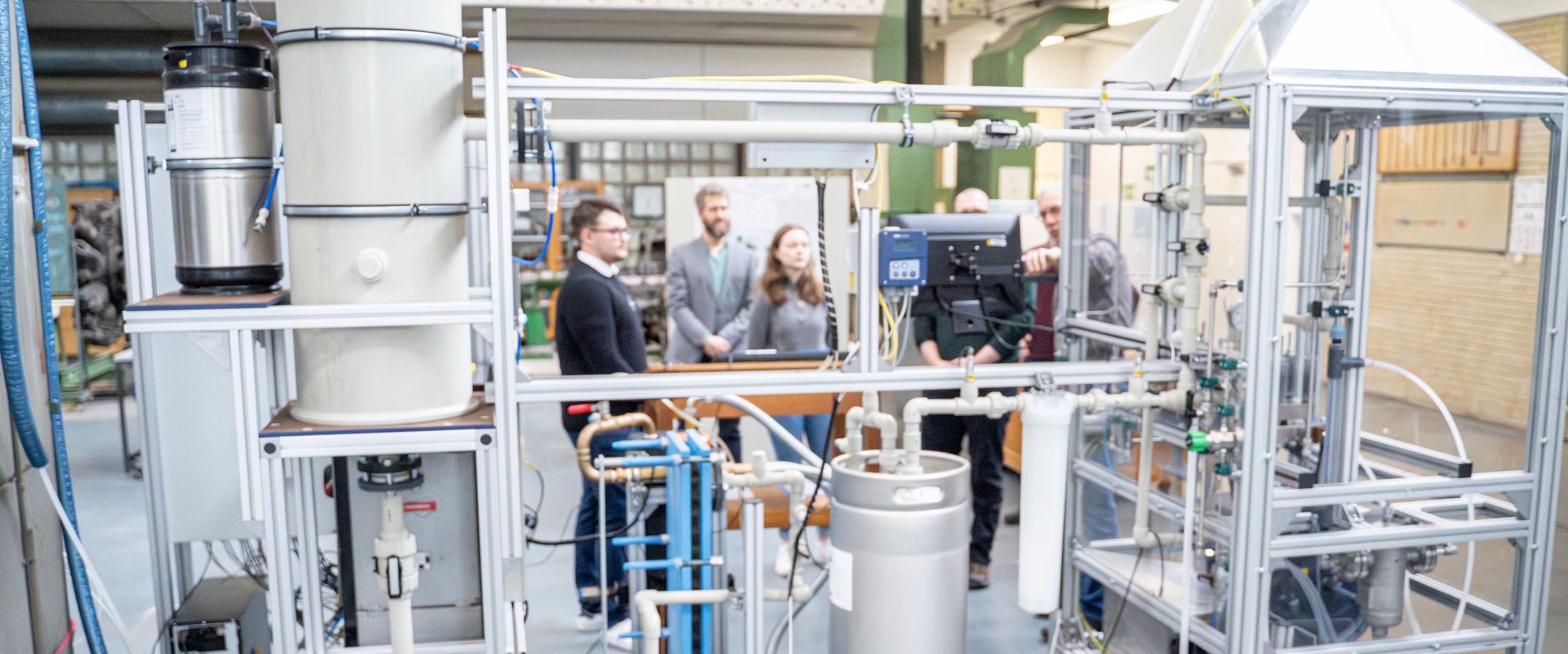 PEM-Großelektrolyseur Das Foto zeigt den Aufbau eines eltrischen gerätes in einer Werkhalle. Dahinter stehen Menschen und betrachten es