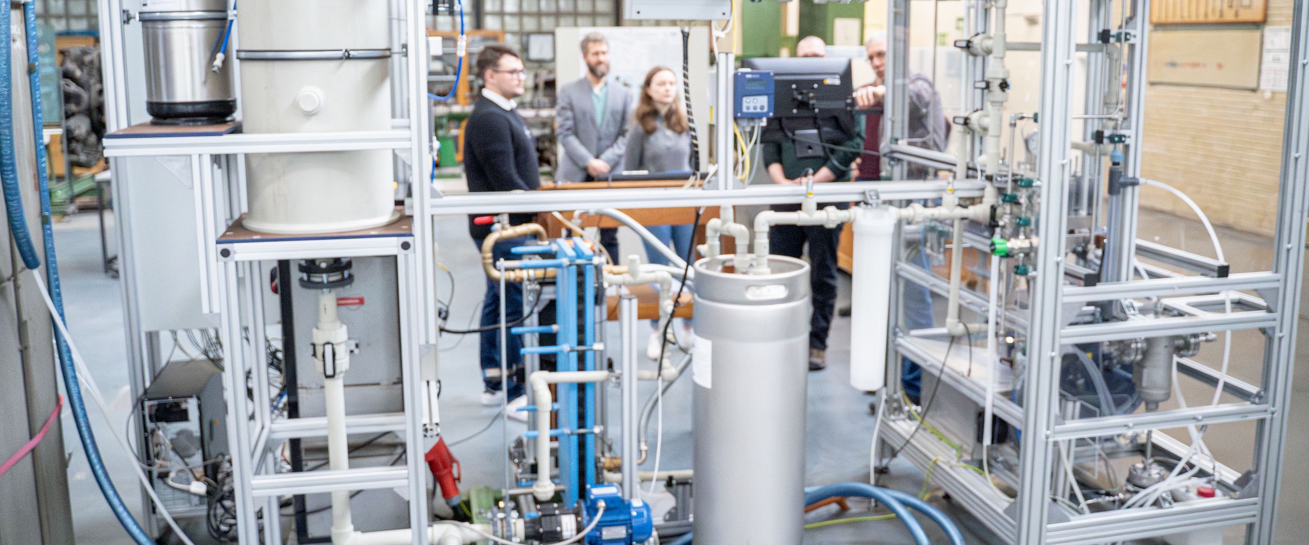 PEM-Großelektrolyseur Das Foto zeigt den Aufbau eines eltrischen gerätes in einer Werkhalle. Dahinter stehen Menschen und betrachten es