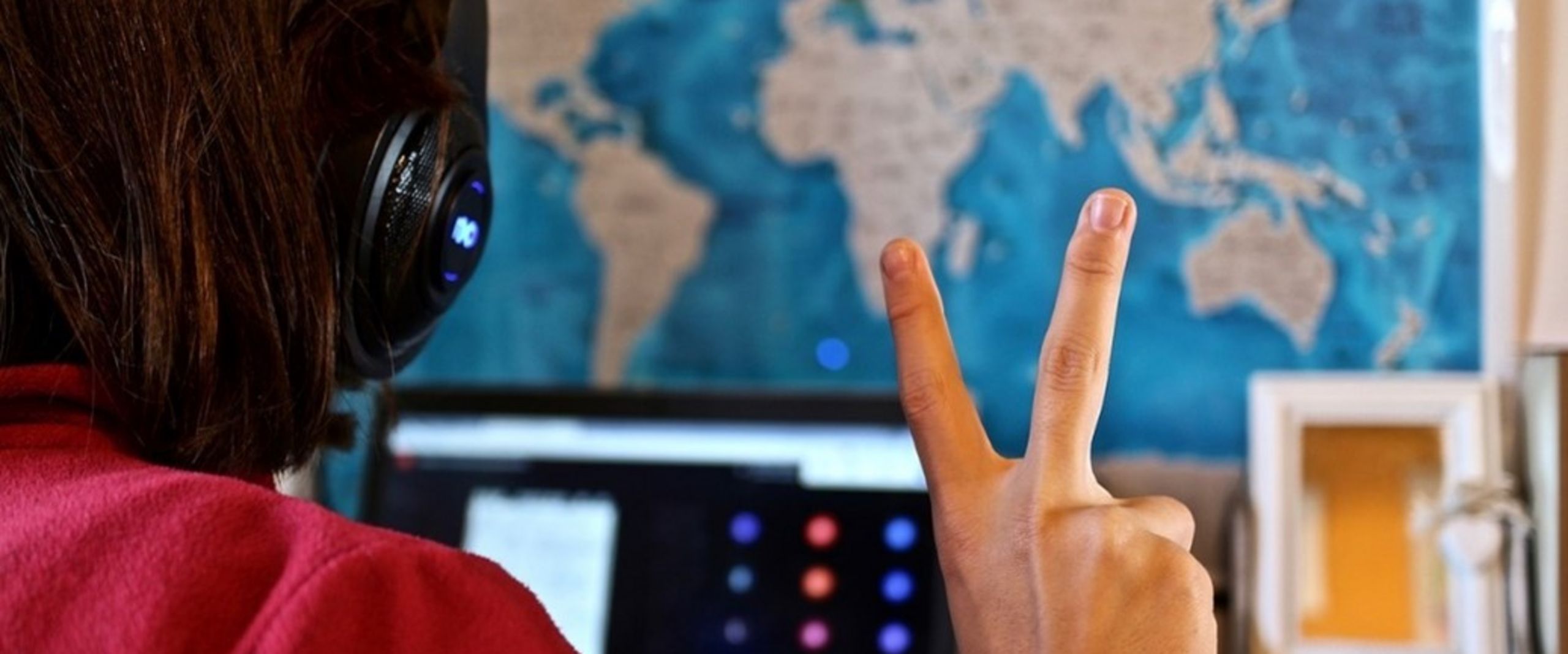 Auf dem Bild ist im Hintergrund eine Weltkarte zu sehen. Im Vordergrund sieht man eine Person von hinten, die Kopfhörer trägt, auf einen Bildschirm schaut und mit der rechten Hand ein Peace-Zeichen darstellt.